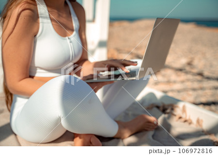 Brown-haired computer sea. A young woman with long hair in white leggings and a top sits on the sand by the sea and uses a laptop 106972816