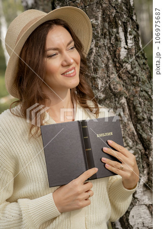 A young beautiful woman in a dress and hat stands and reads a Bible book in nature. Romantic and vintage photo of a beautiful girl. The book has the Bible written on it. Faith in God concept A young beautiful woman in a dress and hat stands and reads a Bible book in nature. Romantic and vintage photo of a beautiful girl. The book has the Bible written on it. Faith in God concept 106975687