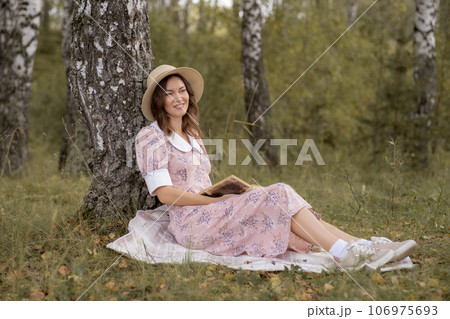 A young beautiful woman in a dress and a round hat reads a book outdoors in the forest and drinks tea. Romantic and vintage photo of a beautiful girl. Reading and relaxation A young beautiful woman in a dress and a round hat reads a book outdoors in the forest and drinks tea. Romantic and vintage photo of a beautiful girl. Reading and relaxation 106975693