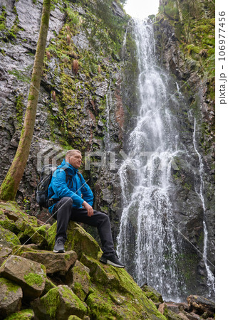 Tourist attraction of Germany - falls of Burgbach Waterfall near Schapbach, Black Forest, Baden-Wurttemberg, Germany. Man hiker in blue jacket standing on stone and looks at flow of falling water 106977456