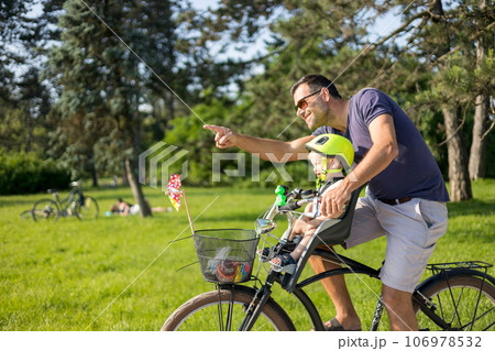 Look over there. Active family day in nature. Father and son ride bike through city park on unny summer day. A cute boy is sitting in front bicycle chair while father rides bicycle. Father son bonding 106978532