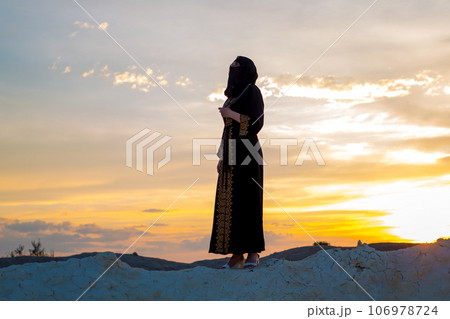 An Islamic woman prays while standing on a mountain at sunset. Religious tradition evening prayer An Islamic woman prays while standing on a mountain at sunset. Religious tradition evening prayer 106978724