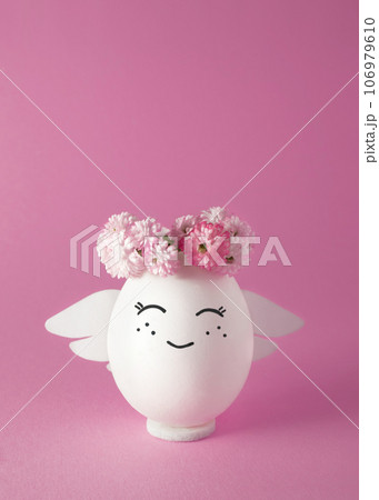 Smiling angel in a wreath of flowers. Easter egg. Pink background, space for text. Smiling angel in a wreath of flowers. Easter egg. Pink background, space for text. 106979610