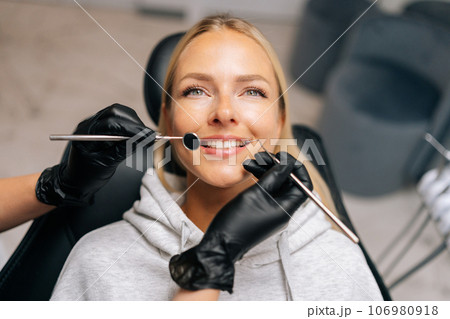 Close up portrait of pretty young woman patient smiling looking away while sitting on dental chair in dentistry clinic. Close-up hands of unrecognizable female dentist in gloves holding dentist tools. 106980918