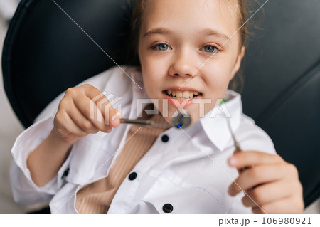 Closeup portrait of cute little child girl holding dentistry mirror and tool lying on dental chair in stomatologist clinic, looking at camera. Concept of children teeth treatment, pediatric checkup. 106980921
