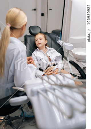 Vertical shot of sad little girl sitting on dental chair and complaining about pain visiting pediatric dentist. Prevention and prophylaxis of dental diseases. Concept of children teeth treatment. 106980922