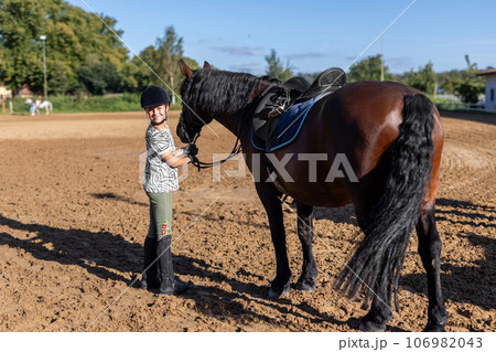 Horse riding school. Little children girls at group training equestrian lessons at outdoors ranch horse riding hall. Cute little beginner blond girl kid near beautiful brown horse 106982043