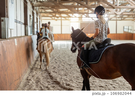 Horse riding school. Little children girls at group training equestrian lessons in indoor ranch horse riding hall. Cute little beginner blond girl kid in helmet sitting on brown horse horseback Horse riding school. Little children girls at group training equestrian lessons in indoor ranch horse riding hall. Cute little beginner blond girl kid in helmet sitting on brown horse horseback 106982044