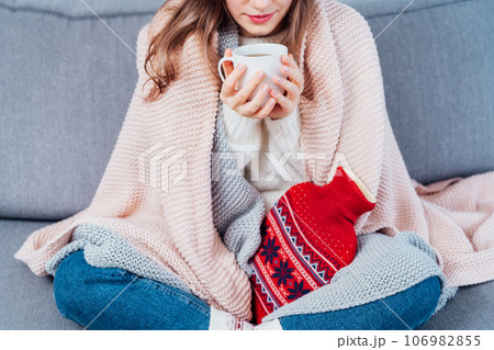 Woman freezes in wintertime. Young girl wearing warm woolen socks and wrapped into two blankets, holding a cup of hot drink and heating pad while sitting on sofa at home. Keep warm. Selective focus Woman freezes in wintertime. Young girl wearing warm woolen socks and wrapped into two blankets, holding a cup of hot drink and heating pad while sitting on sofa at home. Keep warm. Selective focus 106982855