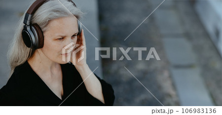 Head shot of a beautiful woman with gray hair sitting on concrete stairs in city, listening to music through wireless headphones. Head shot of a beautiful woman with gray hair sitting on concrete stairs in city, listening to music through wireless headphones. 106983136