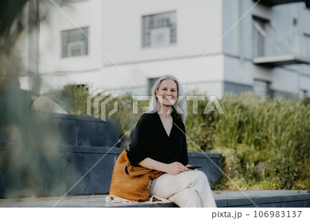 Portrait of a beautiful woman with gray hair sitting on concrete stairs in city, enjoying warm summer weather. 106983137