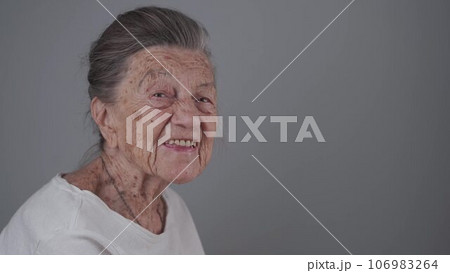 Senior woman 90 years old smiles, laughs and shows a denture on a gray background. Elderly female with deep wrinkles and gray hair happy in a good mood studio portrait looking at camera 106983264