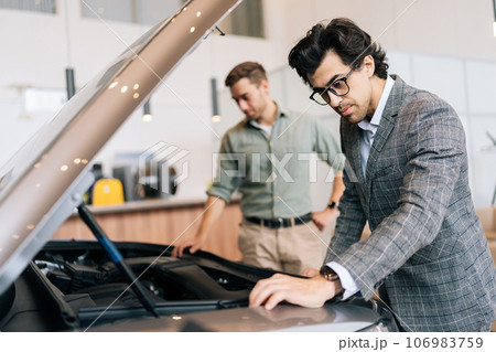 Serious male client visiting dealership office and choosing new car, examine transport engine. Professional salesman describes customer about quality of vehicle in showroom. Concept of buying new auto Serious male client visiting dealership office and choosing new car, examine transport engine. Professional salesman describes customer about quality of vehicle in showroom. Concept of buying new auto 106983759