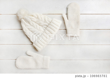 A white knitted hat with a pompom and woolen mittens lie on a white wooden background. Details of winter clothing 106983781