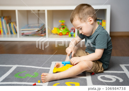 A little toddler boy is playing with a toy helicopter, assembling and disassembling it. Educational toys for kids. Children's room in home. Indoors A little toddler boy is playing with a toy helicopter, assembling and disassembling it. Educational toys for kids. Children's room in home. Indoors 106984738