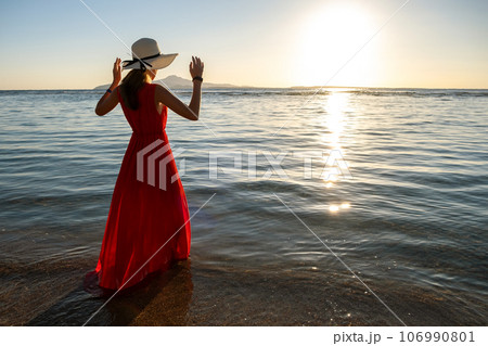 Young woman wearing long red dress and straw hat standing in sea water at the beach enjoying view of rising sun in early summer morning. 106990801