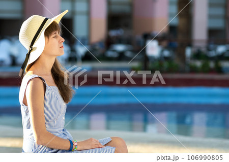Young woman wearing light summer dress and yellow straw hat sitting outside near hotel swimming pool on summer sunny day. 106990805