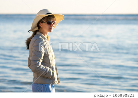 Young woman in straw hat and a dress standing alone on empty sand beach at sea shore. Lonely tourist girl looking at horizon over calm ocean surface on vacation trip. 106990823