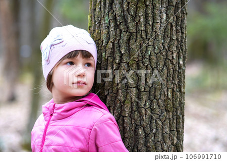 Portrait of young pretty child girl wearing pink jacket and cap leaning to a tree in forest enjoying warm sunny day in early spring outdoors. Portrait of young pretty child girl wearing pink jacket and cap leaning to a tree in forest enjoying warm sunny day in early spring outdoors. 106991710