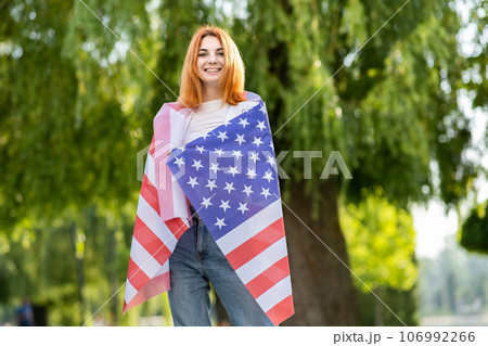 Happy young red haired woman posing with USA national flag on her shoulders standing outdoors in summer park. Pretty girl celebrating United States independence day. Happy young red haired woman posing with USA national flag on her shoulders standing outdoors in summer park. Pretty girl celebrating United States independence day. 106992266