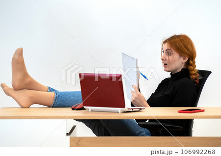 Happy young office worker woman sitting relaxed with feet on table behind working desk with laptop computer, cell phone and notebook. Happy young office worker woman sitting relaxed with feet on table behind working desk with laptop computer, cell phone and notebook. 106992268