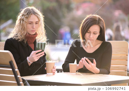 Distracted women browsing their sellphones sitting together at city street cafe. Girlfriends ignoring each other while looking in gadgets. Lack of personal contact between friends in real life concept Distracted women browsing their sellphones sitting together at city street cafe. Girlfriends ignoring each other while looking in gadgets. Lack of personal contact between friends in real life concept 106992741
