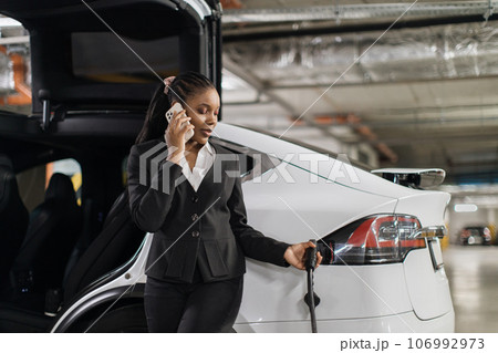 Female with mobile putting charger gun in EV port in garage Female with mobile putting charger gun in EV port in garage 106992973