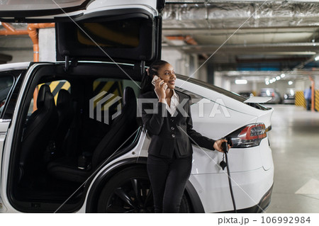 Lady chatting via gadget while charging e-car in parking Lady chatting via gadget while charging e-car in parking 106992984