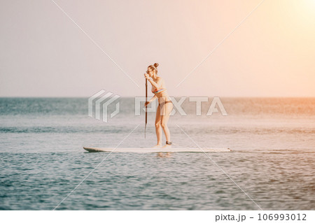 Sea woman sup. Happy woman in a rainbow-coloured bikini stands on SUP board. She is paddling through the calm water, creating ripples along the way. Idyllic sunset. Active lifestyle at sea or river. Sea woman sup. Happy woman in a rainbow-coloured bikini stands on SUP board. She is paddling through the calm water, creating ripples along the way. Idyllic sunset. Active lifestyle at sea or river. 106993012