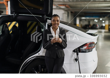 Businesswoman with mobile waiting for e-car being filled up Businesswoman with mobile waiting for e-car being filled up 106993032