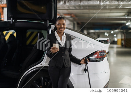 Female with gadget standing near EV in charging session Female with gadget standing near EV in charging session 106993041