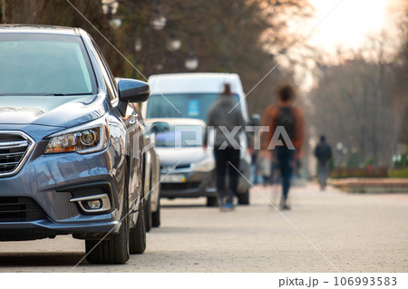 Cars parked in a row on a city street side with blurred people walking on pedestrian zone Cars parked in a row on a city street side with blurred people walking on pedestrian zone 106993583