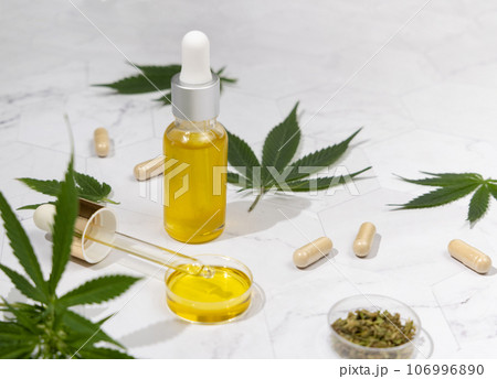 Dropper bottle with CBD oil, pipette and capsules near cannabis leaves close up, hard shadows 106996890