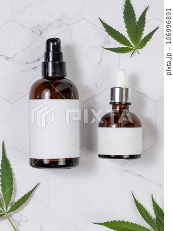 Cosmetic bottles with blank labels near green cannabis leaves on marble table. Mockup Cosmetic bottles with blank labels near green cannabis leaves on marble table. Mockup 106996891