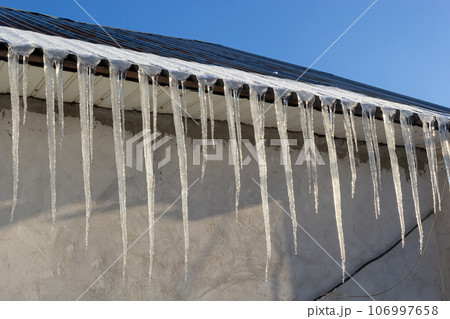 Icicles hang on the roof of a house against a bright blue sky. Spring landscape with icicles hanging from the roof of the house. Set of snow icicles, snow hat Icicles hang on the roof of a house against a bright blue sky. Spring landscape with icicles hanging from the roof of the house. Set of snow icicles, snow hat 106997658
