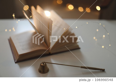 Home autumn composition with book and candle extinguisher in the interior of the room, on a blurred background with a garland. Home autumn composition with book and candle extinguisher in the interior of the room, on a blurred background with a garland. 106998548