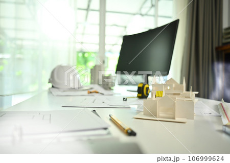 Architect desk with house model, blueprints, safety helmet and computer monitor on white table 106999624