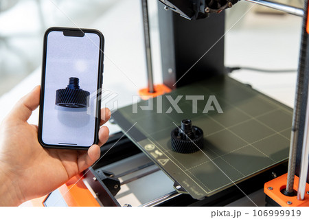 Man holding smartphone with detail on screen, working on 3D printer in creative lab. Man holding smartphone with detail on screen, working on 3D printer in creative lab. 106999919