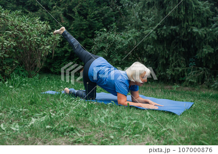 Athletic 50s senior woman doing poses at yoga mat outdoors dressed in sport clothes alone in the park or forest in nature  107000876
