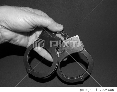 Police handcuffs in a man`s hand, a black-and-white photo on a dark background. 107004686