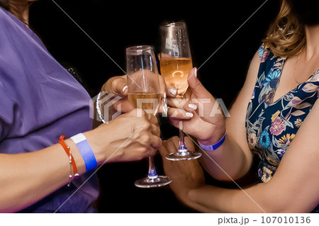 Two adult women clinking glasses of chilled champagne alcohol on a dark background, close-up 107010136