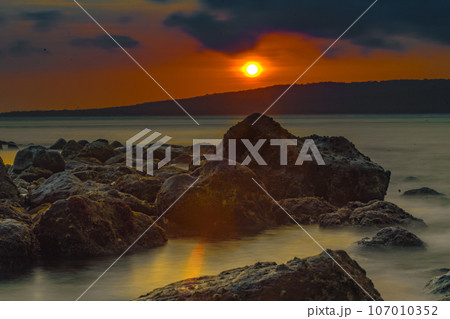 The stunning sunrise view in the Bali Strait, Indonesia, with a foreground of rocky shores along the beach, was captured using the long exposure photography technique. The stunning sunrise view in the Bali Strait, Indonesia, with a foreground of rocky shores along the beach, was captured using the long exposure photography technique. 107010352