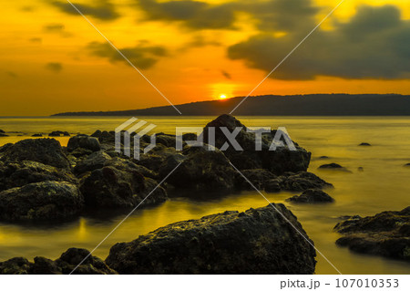 The stunning sunrise view in the Bali Strait, Indonesia, with a foreground of rocky shores along the beach, was captured using the long exposure photography technique. The stunning sunrise view in the Bali Strait, Indonesia, with a foreground of rocky shores along the beach, was captured using the long exposure photography technique. 107010353