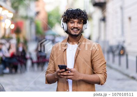 Portrait of a young handsome Indian man standing outside on the street wearing white headphones and holding a phone, smiling at the camera. 107010511