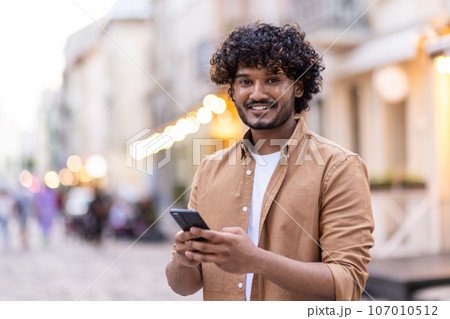 Portrait of a smiling young Indian man traveling tourist standing in the middle of the street, using the phone and smiling at the camera. Portrait of a smiling young Indian man traveling tourist standing in the middle of the street, using the phone and smiling at the camera. 107010512