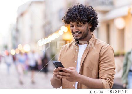 Young smiling Indian man standing outside and using phone, texting, chatting, calling. Young smiling Indian man standing outside and using phone, texting, chatting, calling. 107010513