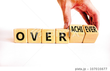 Achiever or overachiever symbol. Businessman turns wooden cubes and changes word Achiever to Overachiever. Beautiful white background. Business achiever or overachiever concept. Copy space. Achiever or overachiever symbol. Businessman turns wooden cubes and changes word Achiever to Overachiever. Beautiful white background. Business achiever or overachiever concept. Copy space. 107010877