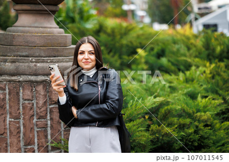 A young pretty girl wearing a black leather jacket walks down the street in a park and uses her smartphone for work. The work of a freelancer and blogger. A young pretty girl wearing a black leather jacket walks down the street in a park and uses her smartphone for work. The work of a freelancer and blogger. 107011545