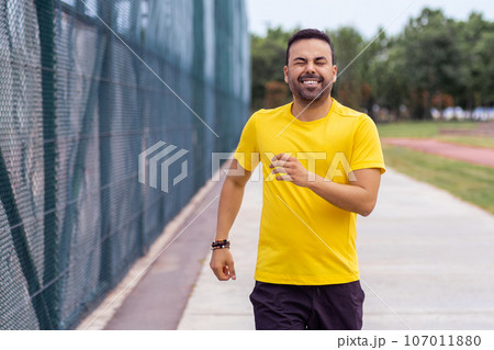Energetic man in a yellow t-shirt goes for a jog in a city park arena on a sunny summer day, dedicated to his training. Energetic man in a yellow t-shirt goes for a jog in a city park arena on a sunny summer day, dedicated to his training. 107011880