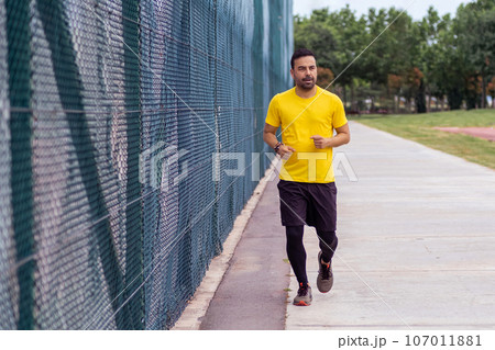 Health-conscious man, wearing a comfortable tracksuit, runs along a road beside a high mesh fence on a sports ground in an urban park, actively pursuing fitness and body development. Health-conscious man, wearing a comfortable tracksuit, runs along a road beside a high mesh fence on a sports ground in an urban park, actively pursuing fitness and body development. 107011881